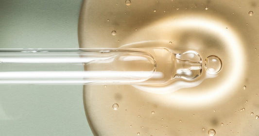 A serum dropper releasing a golden drop into a pool of skincare serum on a soft sage background